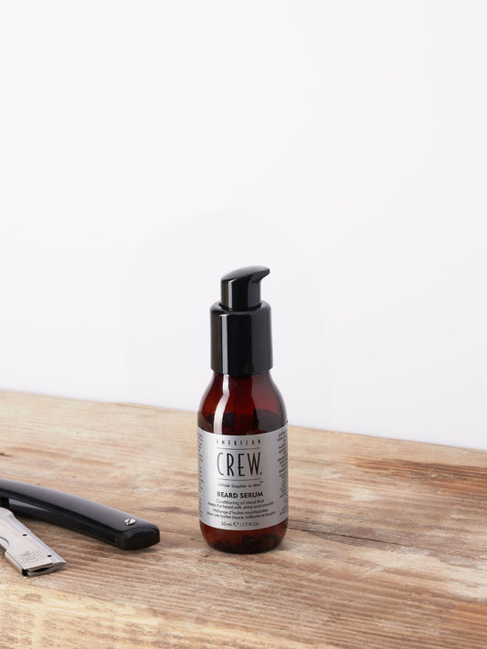 American Crew Beard Serum 50ml — 50 ml pump bottle standing on a wooden barber counter beside a straight razor