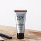 American Crew 2 In 1 Skin Moisturizer & Beard Conditioner — 100 ml tube standing on a wooden barber counter beside a straight razor