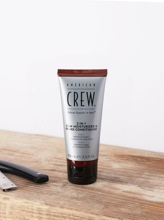 American Crew 2 In 1 Skin Moisturizer & Beard Conditioner — 100 ml tube standing on a wooden barber counter beside a straight razor