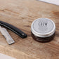 American Crew Beard Balm 62ml — 62 ml balm tin standing on a wooden barber counter beside a straight razor