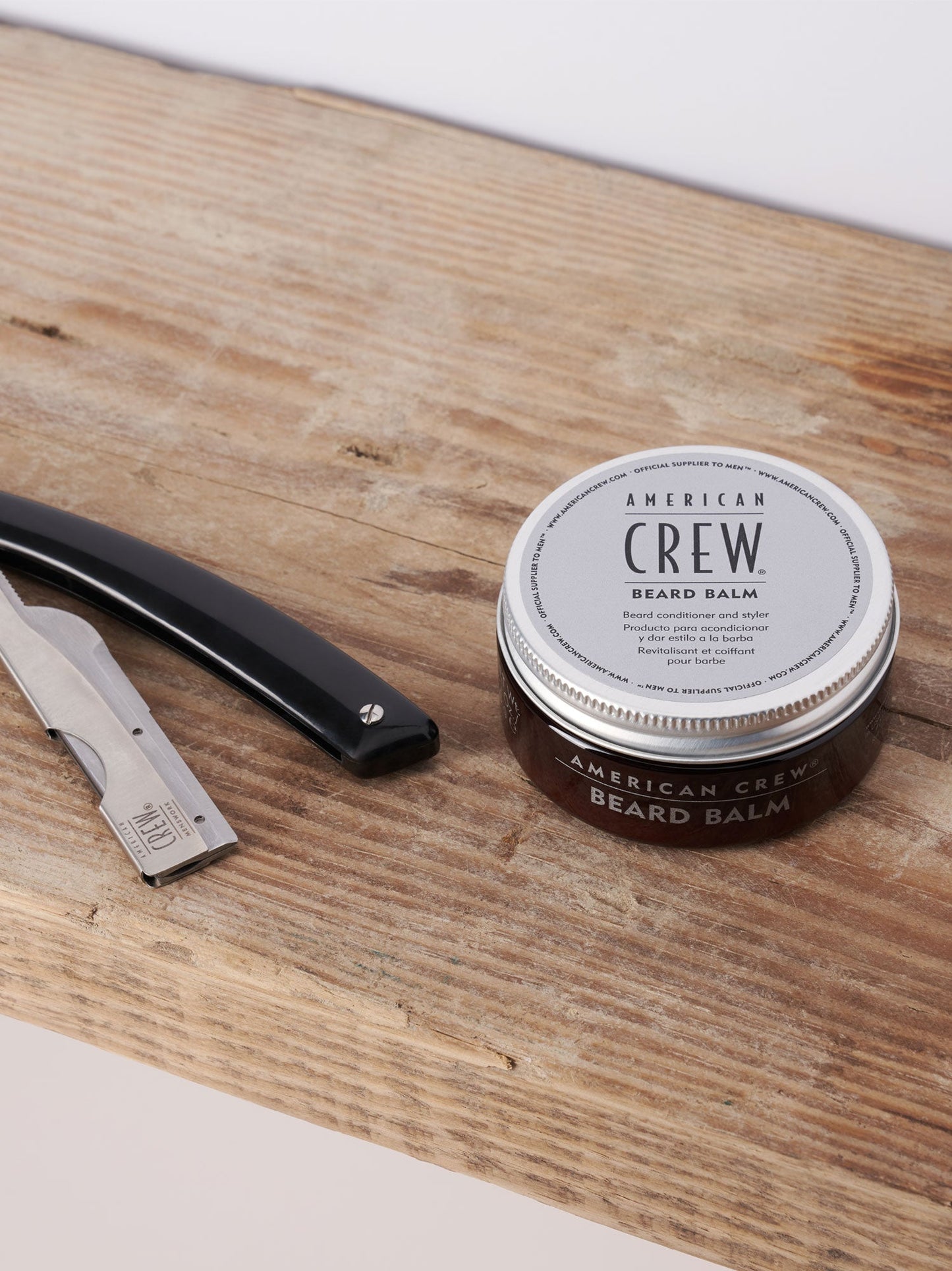 American Crew Beard Balm 62ml — 62 ml balm tin standing on a wooden barber counter beside a straight razor