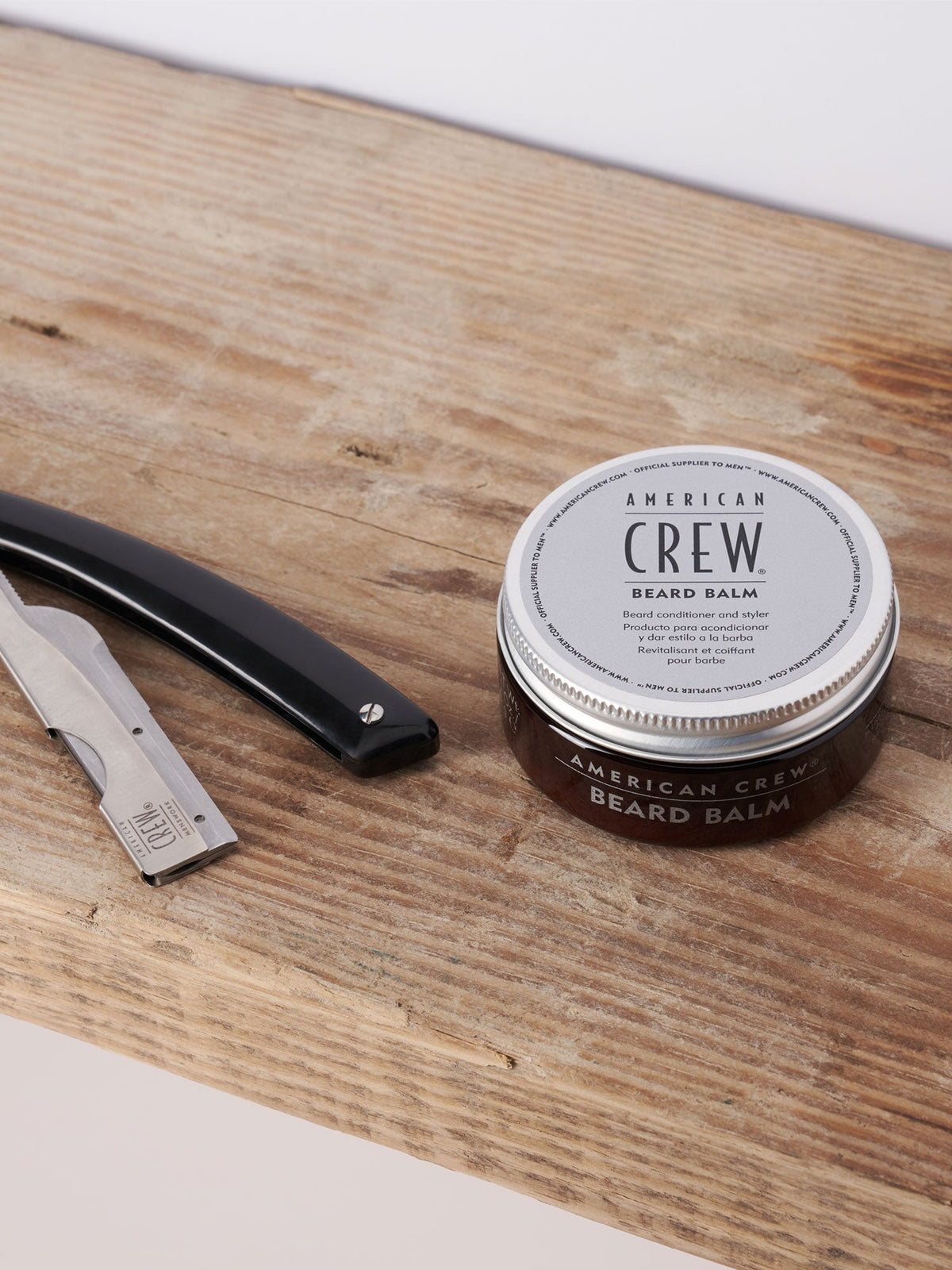 American Crew Beard Balm 62ml — 62 ml balm tin standing on a wooden barber counter beside a straight razor