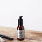 American Crew Beard Serum 50ml — 50 ml pump bottle standing on a wooden barber counter beside a straight razor